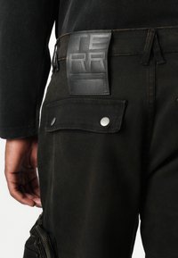 Zwarte stoffen broek met een gestructureerd leren label met de tekst "TERRA" op de tailleband, voorzien van een zijrits zak en metalen drukknoopdetails.