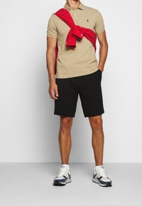 Beżowa koszulka polo z zielonym logo, czarne szorty i czerwona kurtka związana na ramionach. Szare i granatowe sneakersy dopełniają strój.