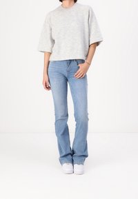 Maglione a maglia grigio chiaro maniche corte abbinato a jeans azzurri a zampa d'elefante. Il modello indossa sneakers bianche, in piedi su uno sfondo semplice.