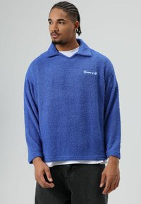 Blauwe fleece trui met een kraag, lange mouwen en een gestructureerde stof. Voorzien van een wit logo borduursel op de borst en een losse pasvorm.