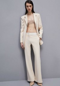 Traje beige a medida con pantalones de pierna ancha y una parte superior ajustada, hecho de tejido suave. Incluye una chaqueta de un solo botón y detalles de costura sutiles.