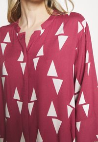 Rödblommig blus med V-ringning, långa ärmar och ett vitt triangulärt mönster. Tillverkad av ett mjukt, lätt tyg med en avslappnad passform.