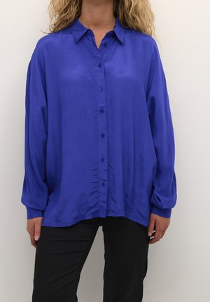 Osoba ubrana w luźną, długą, kobaltowo-niebieską koszulę na guziki i czarne spodnie, stojąca na jasnym, jednolitym tle.