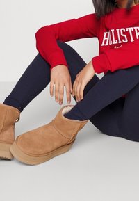 Botas de ante marrón hasta el tobillo con forro de borreguillo, tacón bajo y punta redondeada, combinadas con leggings oscuros y una sudadera roja.
