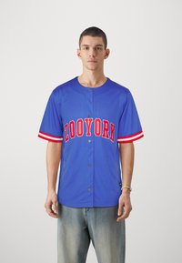 Blå trøye med røde detaljer og teksten "ZOONYORK". Korte ermer med stripete mansjetter. Frontknapper og myk stoff. Brukt med lyse jeans.