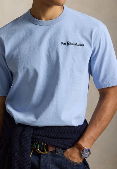 Ανοιχτό μπλε βαμβακερό T-shirt με κοντά μανίκια, στρογγυλή λαιμόκοψη και κεντημένο λογότυπο. Φορεμένο με σκούρο πλεκτό πουλόβερ δεμένο γύρω από τη μέση.