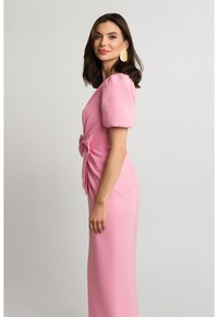 Różowa sukienka z krótkimi bufkami, akcentem w postaci kokardy z boku oraz smukłym, wydłużonym krojem. Gładki materiał i proste linie tworzą elegancki wygląd.