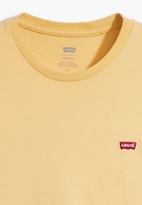 Żółta bawełniana koszulka z okrągłym dekoltem, z małą czerwoną łatą z logo Levi's po lewej stronie. Gładka faktura, minimalistyczny design.