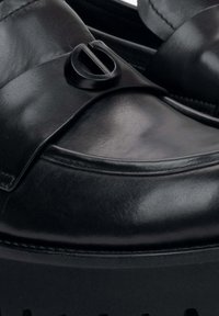 Czarne skórzane buty wsuwane z okrągłym noskiem, wyraźnym paskiem i okrągłym logo na cholewce. Teksturowana, gładka powierzchnia.