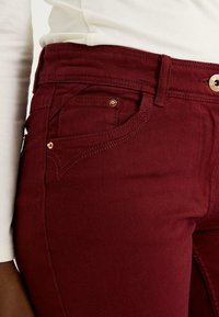 Jeans burdeos con un diseño ajustado, dos bolsillos frontales, detalles en hardware en tono dorado y costuras detalladas a lo largo de las costuras.