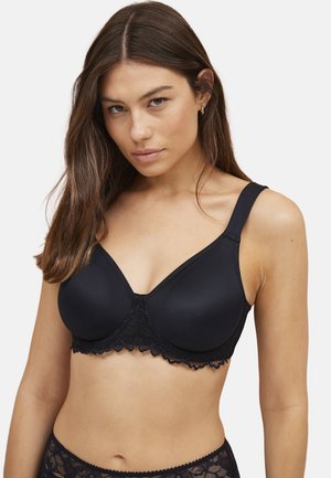 Kvinde iført en sort bh med blonder og matchende blonderundertøj, med langt brunt hår og et neutralt ansigtsudtryk, på en ensfarvet baggrund.