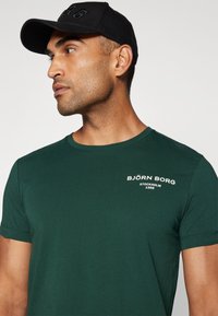 Grønn kortermet t-skjorte i mykt stoff med rund hals og hvit trykt logo på brystet. Svart caps med preget tekst.