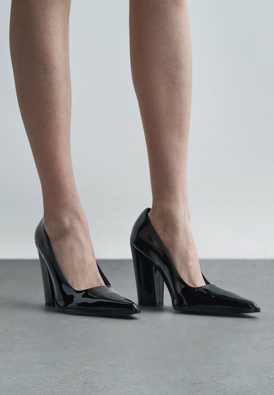 Μαύρα παπούτσια γυαλιστερής πατέντας με ψηλό τακούνι και μυτερή μύτη. Λείος, γυαλιστερός επιφάνεια με καθαρό, κομψό σχέδιο.
