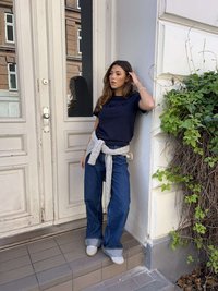 Tengerészkék póló "love" felirattal, magas derekú kék farmer, szürke pulóver derékban megkötve, fehér tornacipő, egy ajtó mellett állva, zöld növényzettel.