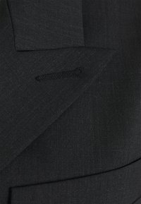 Blazer in tessuto nero con superficie liscia e strutturata, leggermente lucida. Dettagli notevoli includono una piccola tasca cucita e rever affilati.