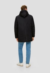 Schwarze gepolsterte Jacke mit Kapuze, die eine elegante, glatte Textur und einen langen Schnitt aufweist, kombiniert mit hellblauen Jeans und weißen Sneakers.