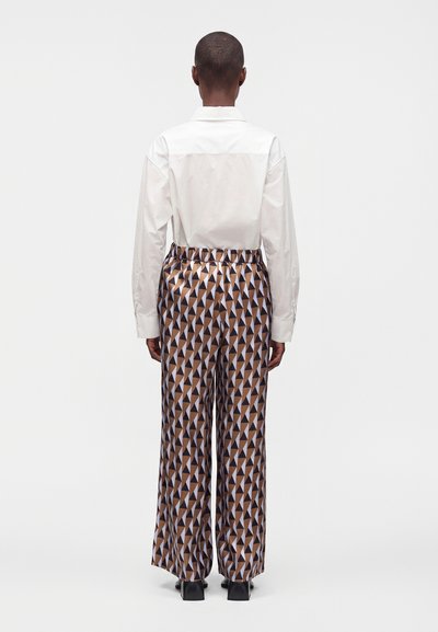 Personne de dos portant une chemise blanche à manches longues et un pantalon large marron à motif géométrique avec des chaussures noires.