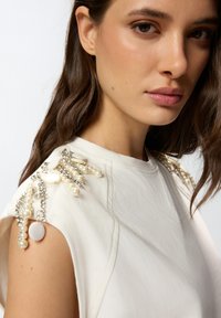 Top blanco con detalles decorativos en el hombro, adornado con perlas, cuentas y piedras transparentes. Tejido suave con un ligero brillo y forma estructurada.