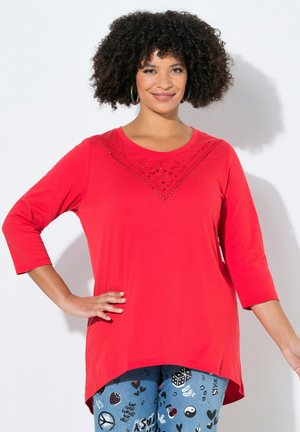 Kvinde med krøllet hår iført en rød langærmet top med dekorative nitter og høj-lav kant, kombineret med blå mønstrede jeans.
