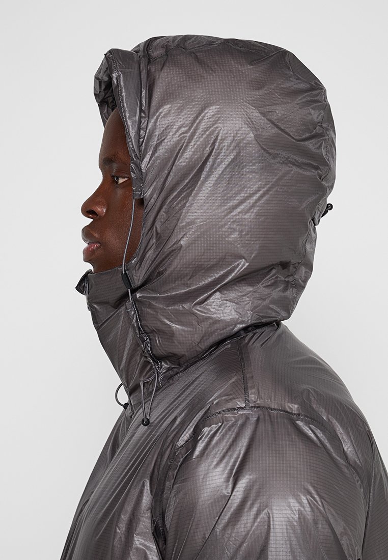 Persona in giacca grigia, lucida, con cappuccio vista di lato, con il cappuccio che copre completamente testa e collo.