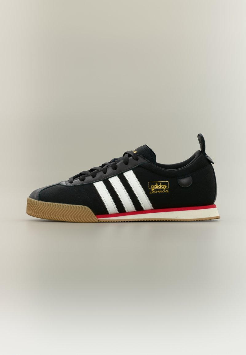 Black Adidas Samba sneaker with white stripes, gum sole, red midsole accent, and gold logo on a neutral background.