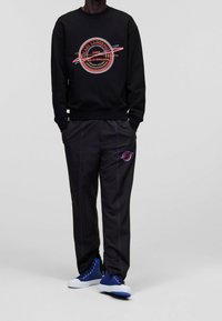 Felpa nera con stampa grafica colorata, abbinata a pantaloni neri con cuciture viola e tasche laterali, completata da sneaker blu.