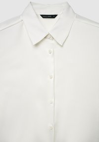 Camicia bianca a maniche lunghe con bottoni e colletto a punta, con etichetta nera della taglia "CALLIOPE · S" all'interno del colletto.