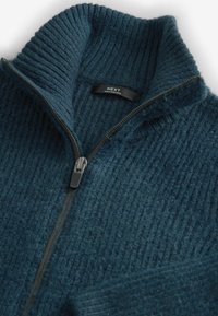 Maglione zip-up a coste di un blu scuro, con colletto alto, dotato di una zip argentata e un'etichetta "NEXT" cucita all'interno.