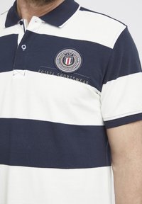 Tmavo modré pruhované polo tričko s golierom, so vyšitým kruhovým logom a textom "1924 SPORTSWEAR".