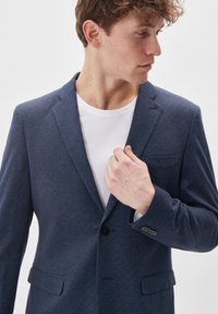 Navy blue blazer with a textured fabric, single button, notched lapels, and a chest pocket, worn over a white T-shirt.