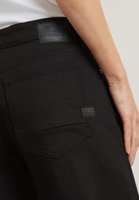 Jeans din denim negru prezintă o etichetă din piele cu branding, un buzunar din spate cu detalii de cusătură și o textură netedă.