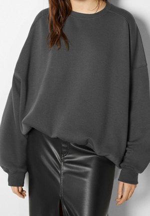 Kobieta w za dużej, ciemnoszarej bluzie i czarnej skórzanej spódnicy z rozcięciem z przodu, pozująca na tle gładkiej ściany.