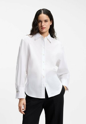 Frau mit dunklem welligem Haar, die ein weißes langärmeliges Hemd und schwarze Hose trägt, steht mit einer Hand in der Tasche.