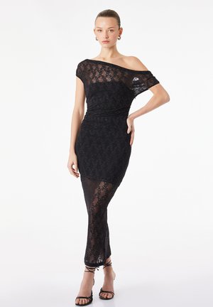 Kobieta ubrana w dopasowaną, przejrzystą czarną koronkową sukienkę z jednym odsłoniętym ramieniem oraz czarne, wysokie sandały na obcasie z paskami, stojąca z ręką na biodrze.