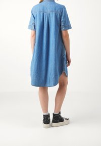 Abito camicia in denim con maniche corte, patta con bottoni e spacchi laterali. Indossato con sneakers alte nere. Colore azzurro chiaro.