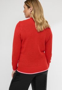 Czerwony sweter o teksturowanym splocie, z długimi rękawami i ściągaczem na dole. Udekorowany różową paskiem na dole. Noszony z czarnymi spodniami pokrytymi cekinami.