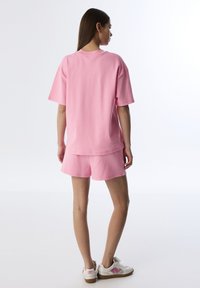 Mujer de pie, vistiendo una camiseta rosa holgada y pantalones cortos a juego, zapatillas blancas con acentos rosas, frente a un fondo blanco liso.