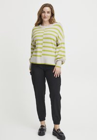 Gestreifter Pullover in hellem Beige mit limettengrünen Streifen, entspannter Schnitt, geripptem Saum. Kombiniert mit schwarzen, taillierten Hosen und schwarzen Slippern.