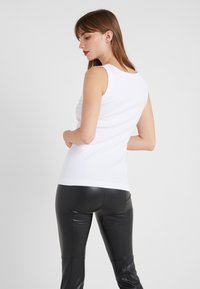 Witte mouwloze top met een geribbelde textuur, gecombineerd met zwarte nepleer broeken. De pasvorm is nauwsluitend, waardoor de taille en de silhouet goed tot zijn recht komen.