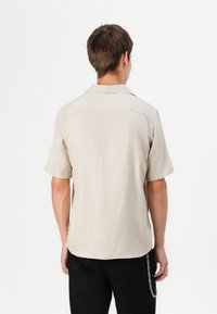 Short-sleeve beige shirt with a straight cut, open collar, and smooth texture. Features a double panel design at the back.