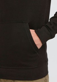 Schwarzer Hoodie aus strukturiertem Stoff, mit einer Fronttasche und gerippten Bündchen. Nahtdetails entlang dem Saum und den Taschenkanten.