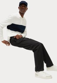 Polo a maniche lunghe a righe bianche e navy, pantaloni neri e sneakers bianche. Modello seduto, che indossa un cappellino navy. Texture del tessuto morbide.