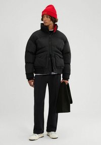 Czarna kurtka puchowa z wysokim kołnierzem i zamek błyskawicznym z przodu, z ribbedmanżetami, nałożona na białą koszulę, w połączeniu z ciemnymi spodniami i czerwoną czapką beanie.