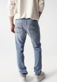 Jeans in denim azzurro chiaro con vestibilità slim, caratterizzati da un lavaggio sbiadito, doppie tasche posteriori e dettagli di cucitura sottili. Indossati con una maglia color crema.