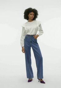 Blusa branca de manga longa com detalhes de volantes, combinada com calças de ganga azuis de cintura alta. A modelo usa sapatos de salto agudo roxos. Fundo simples.
