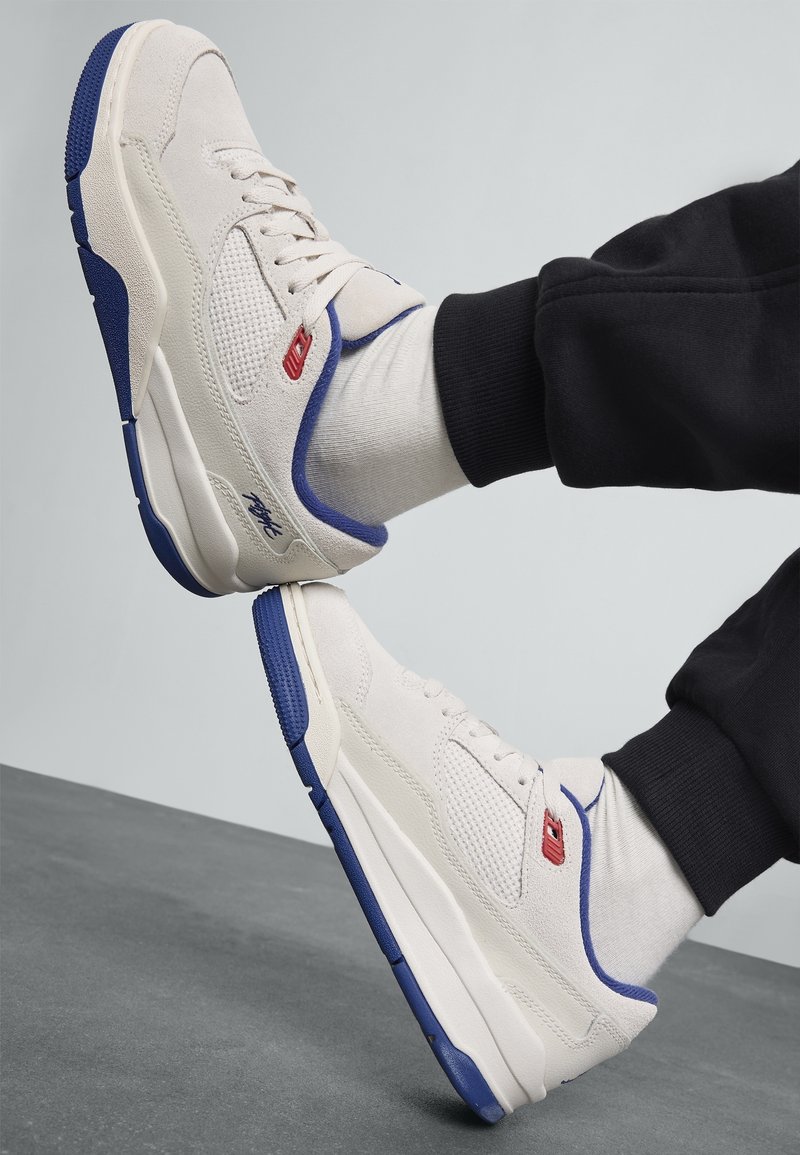 Baskets blanches et bleues portées avec des chaussettes blanches et un pantalon noir, reposant sur un sol gris avec un fond gris clair.