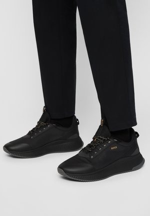 Black low-top sneakers with textured sides, branded "BOSS" in gold, worn with black pants on a light gray background.