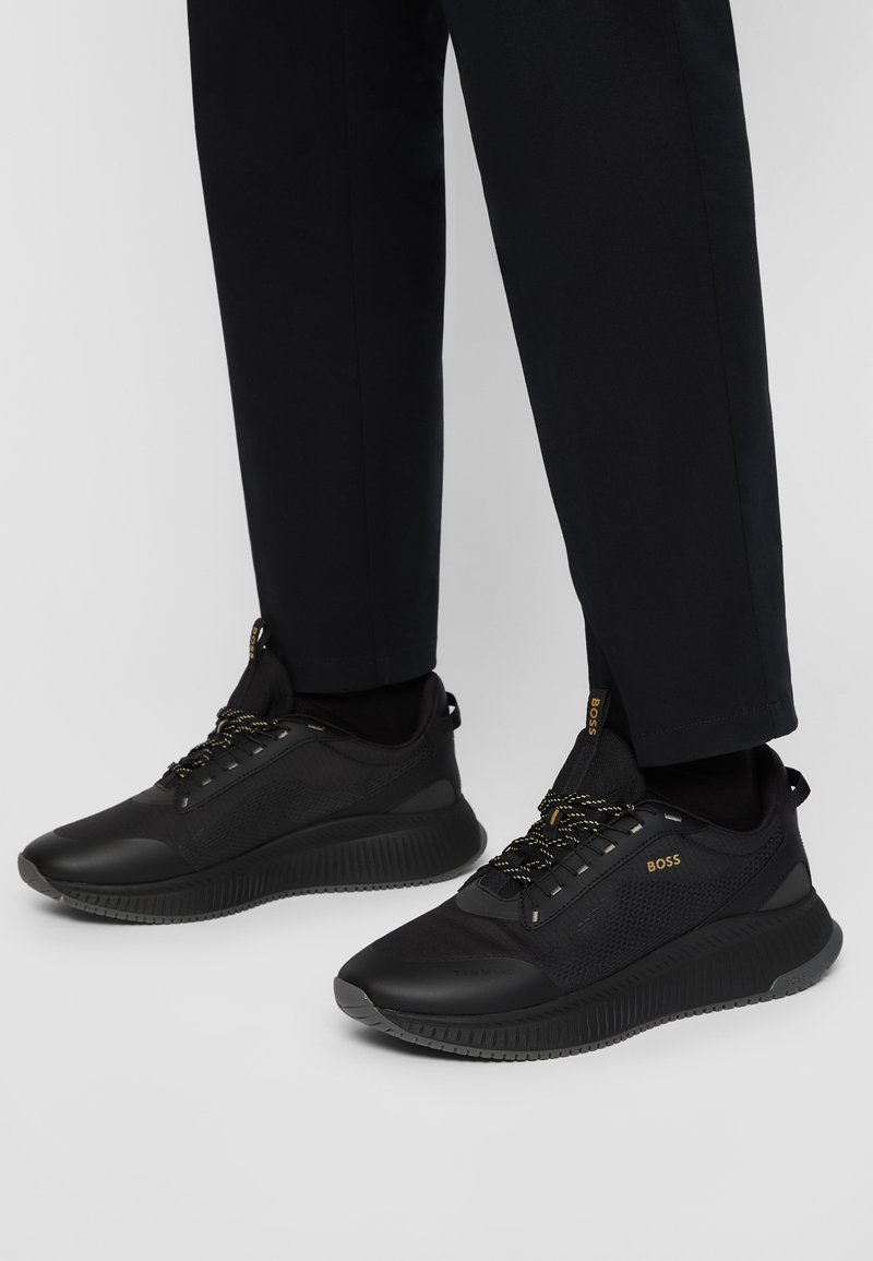 Baskets basses noires avec côtés texturés, estampillées "BOSS" en or, portées avec un pantalon noir sur fond gris clair.