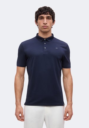 Uomo che indossa una polo blu navy aderente a maniche corte con bottoni e pantaloni bianchi, in piedi davanti a uno sfondo chiaro e uniforme.