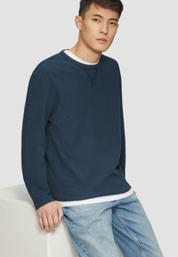 Marineblaues langärmliges Sweatshirt aus weichem Stoff, mit Rundhalsausschnitt und gerippten Bündchen, über einem weißen T-Shirt getragen und mit hellen Jeans kombiniert.
